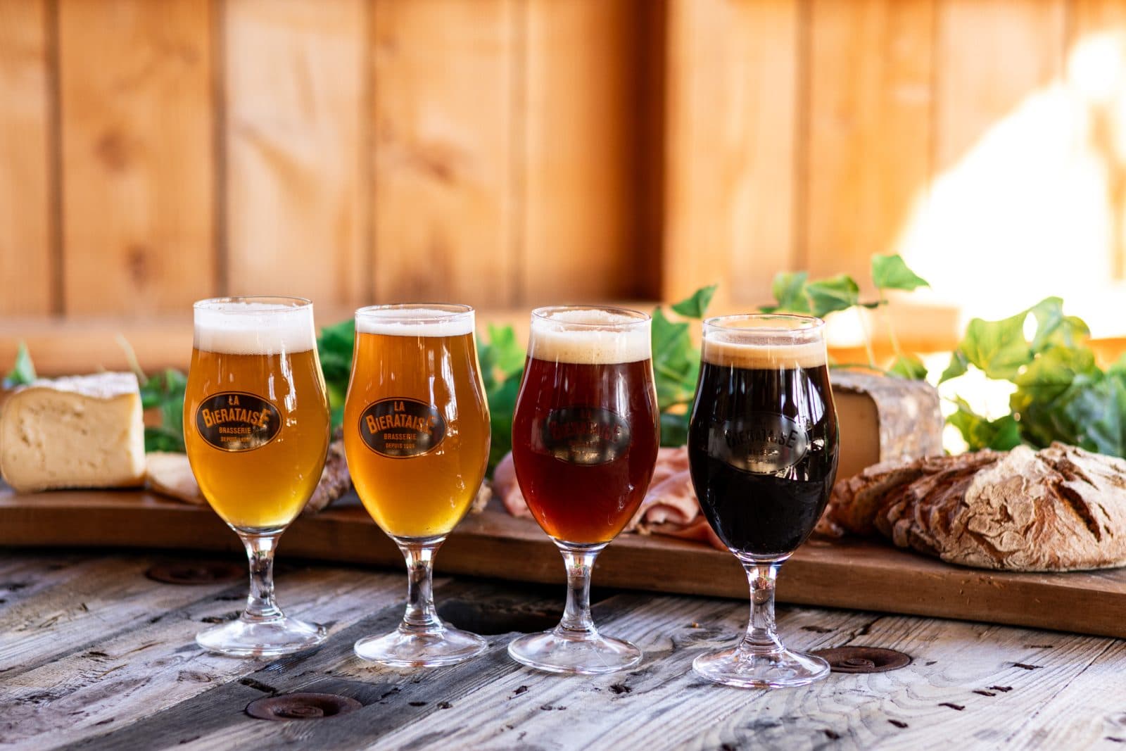 Quatre verres de bière de La Bièrataise, du blond au brun, sur une table en bois avec fromage et pain